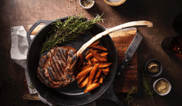 A cast iron skillet with a tomahawk steak, steak fried potatoes and asparagus